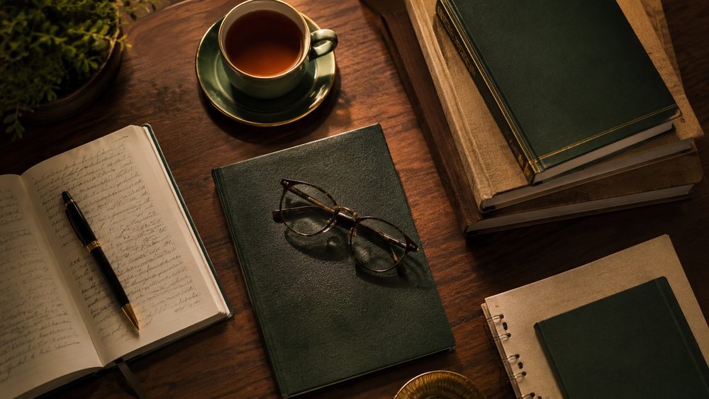 A flat-lay of distance MBA study materials — bound books, a handwritten notebook, reading glasses and a cup of tea on a wooden desk